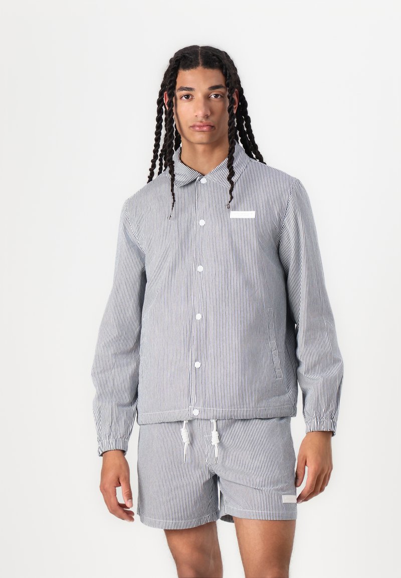 Striped fabric jacket with long sleeves and button closure, paired with matching shorts. Colors are navy and white. Features pockets and drawstrings.