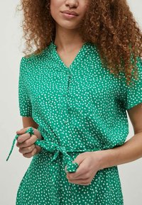 Grön klänning med knappar och vita prickar, korta ärmar, en ficka på vänster sida och ett knytbälte i midjan. Slät tygtextur.