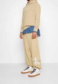 Beige hoodie met logo, gelaagd over een denim shirt. Relaxed-fit beige broek en witte sneakers met marineblauwe accenten en gumzolen, die comfort benadrukken.