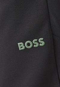 Črn material z teksturiranim, mrežastim vzorcem logotipa "BOSS" v svetlo zeleni barvi, ki je sijoče postavljen na materialu.