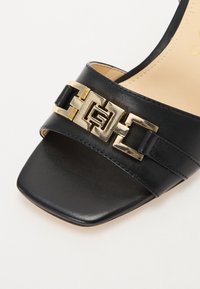 Zwarte leren slide sandaal met een vierkante neus. Voorzien van een gouden decoratieve gesp en een gladde textuur. Eenvoudig ontwerp met kleine accentdetails.