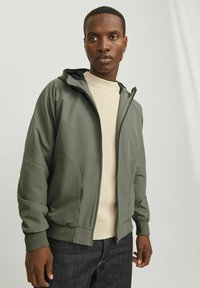 Chaqueta de cremallera verde oliva con capucha, hecha de una tela suave; presenta bolsillos laterales y puños acanalados. Usada sobre un suéter de color beige claro.