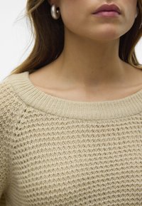Beżowy sweter dzianinowy z szerokim, żebrowanym dekoltem. Teksturowany wzór materiału i dopasowany krój. Widoczne duże srebrne kolczyki w kształcie kół.
