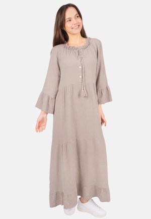 Femme vêtue d'une robe longue beige texturée avec des poignets et un ourlet à volants, trois boutons, et des baskets blanches, souriante et regardant légèrement sur le côté.