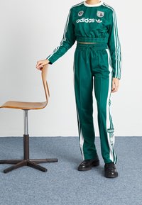 Grön adidas träningsoverall med en kort topp och byxor med elastisk midja samt vita sidostråk. Svarta skor och en trämöbel i bakgrunden.