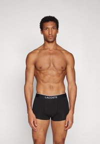 Black cotton boxer shorts with a white Lacoste logo on the waistband, featuring a fitted design and smooth texture.
