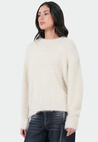 Crème-kleurige trui van zacht, fluffy materiaal, met een ronde halslijn en geribde zoom. Gekoppeld aan donkere spijkerbroek.