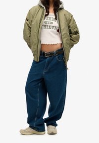 Groene bomberjack met een grijze capuchon, witte cropped top, blauwe baggy jeans en beige sneakers. Opvallende details: zwarte riem met studs.