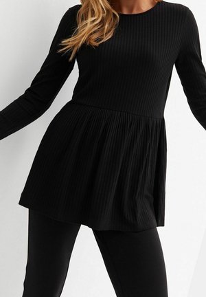 Kobieta ubrana w czarny, prążkowany, długi top peplum z długim rękawem, zestawiony z czarnymi dopasowanymi spodniami na jasnym, jednolitym tle.