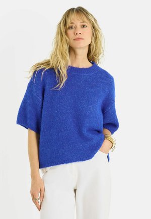 Mujer con cabello largo y rubio, que lleva un suéter azul brillante de manga corta y pantalones blancos, posando con una mano en el bolsillo y pulseras doradas.