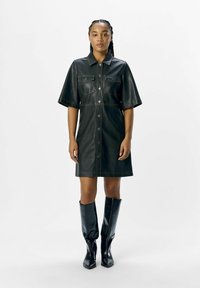 Zwarte leren shirtdress met korte mouwen, knopenluiting aan de voorkant en twee borstzakken. Gestyled met kniehoge zwarte laarzen.
