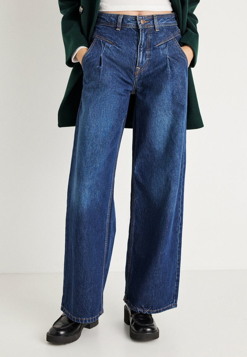 Calças de ganga azuis de perna larga com uma cintura alta, apresentando pregas frontais e bolsos laterais. Bainhas cosidas e uma textura ligeiramente desgastada.