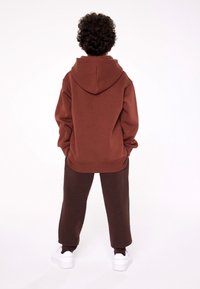 Felpa marrone con cappuccio regolabile e polsini a costine, abbinata a pantaloni della tuta marroni scuri. Scarpe da ginnastica bianche completano l'outfit.