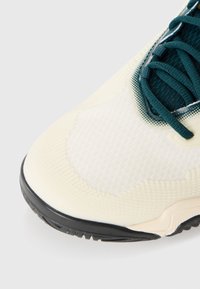 Close-up van een witte sneaker met een gestructureerde neus, donkergroene veters en een zwarte zool op een witte achtergrond.