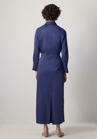 Abito lungo a maniche lunghe blu navy con colletto, cintura e polsini con bottoni. Presenta uno spacco posteriore per facilitare i movimenti e un tessuto testurizzato.