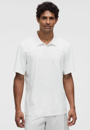 Uomo con capelli corti e ricci che indossa una polo bianca semplice a maniche corte e pantaloni bianchi, in piedi davanti a uno sfondo grigio chiaro.