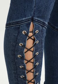 Džínové kalhoty v tmavě modré barvě se zdobným šněrovacím detailem podél dolní části stehna, s očky a křížovým černým šněrováním.
