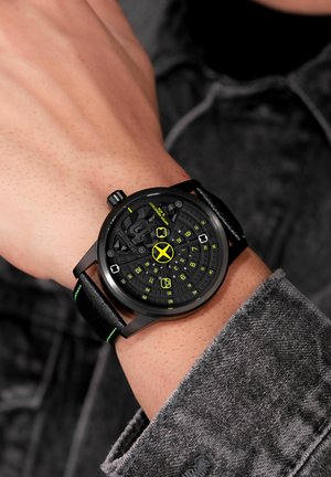 Black wristwatch with a textured dial, neon yellow accents, and a black leather strap with a green stitch. Bold, modern design with a circular shape.