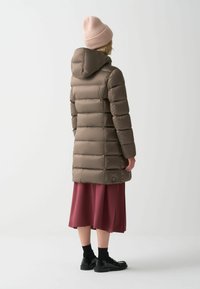 Pufferjas in zacht taupe, met horizontale stiksels, een capuchon en lange mouwen. Draagt over een bordeauxrok en gecombineerd met zwarte schoenen.