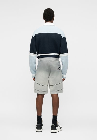 Άνδρας με την πλάτη, φοράει μπλε σκούρο και ανοιχτό μακρυμάνικο πουκάμισο, γκρι σορτς με λογότυπο στην πίσω τσέπη, μαύρες κάλτσες και ασπρόμαυρα αθλητικά παπούτσια.