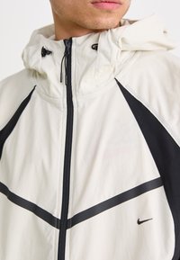 Hvit hettejakke med svarte detaljer, frontglidelås og horisontale striper. Materialet ser lett ut med en jevn tekstur. Nike-logoen er synlig.