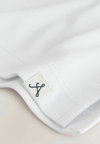Gros plan sur un tissu blanc avec un bord plié et une petite étiquette blanche portant un symbole bouclé sombre brodé près de la couture.