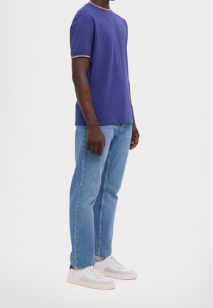 Uomo in piedi che indossa una t-shirt blu a maniche corte, jeans azzurri e scarpe da ginnastica bianche basse su uno sfondo semplice.