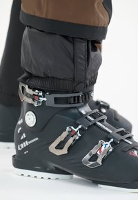 Skischoenen van zwart plastic met verstelbare metalen gespen en een gladde textuur. Voorzien van een rubberen zool met witte accenten en ontwerpdetails.