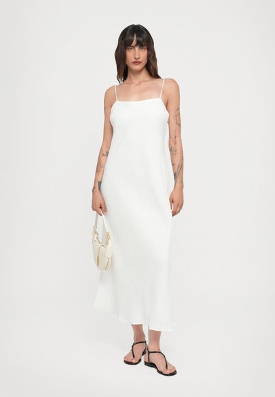 Vestido de lino blanco sin mangas con tirantes finos, de silueta fluida y largo midi. Combinado con sandalias negras y un pequeño bolso blanco.