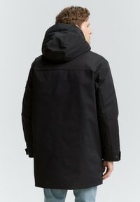 Schwarze Parka mit Kapuze und matter Oberfläche, mit einem langen Schnitt, versteckten Reißverschlüssen und verstellbaren Bündchen. Trägt man über hellblauen Jeans.