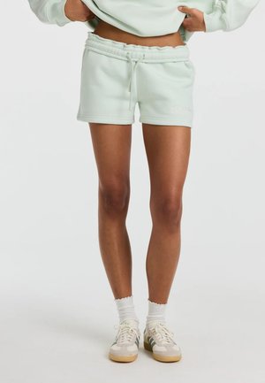 Person iført lysegrønne shorts med snøre, hvide sokker og beige sneakers med grønne striber, stående mod en ensfarvet baggrund.