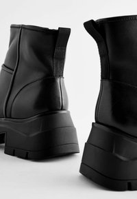 Bottes noires jusqu'à la cheville avec un dessus en cuir synthétique, une semelle épaisse, des coutures proéminentes et des panneaux latéraux extensibles pour un enfilage facile.