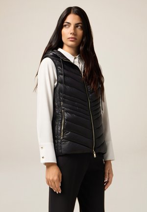 Zwarte puffer vest met een capuchon, voorzien van een chevronpatroon, twee ritszakken en een voorrits. Gedragen over een witte blouse.