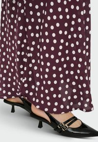Robe midi bordeaux à pois avec un design fluide et une paire d'escarpins noirs en cuir verni, à talons bas, avec des brides autour de la cheville et de petites boucles.