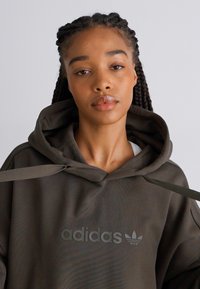 Felpa con cappuccio verde scuro con un grande logo adidas tono su tono sul davanti. Presenta un cappuccio con cordino e una vestibilità rilassata. Tessuto morbido e testurizzato.