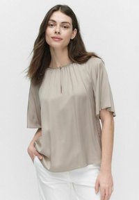 Beige blus med nyckelhålsöppning, korta volangärmar och mjukt, slätt tyg; matchad med vita byxor.