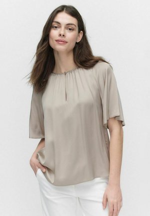 Beige pusero, jossa on avonainen pääntie, lyhyet hulmuavat hihat ja pehmeä, sileä kangas; yhdistetty valkoisiin housuihin.