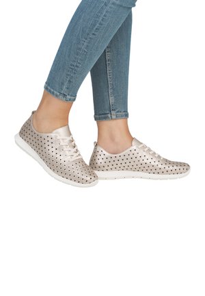 Lekkie sneakers wykonane z metalicznego srebrnego materiału z okrągłymi wycięciami, białą podeszwą i sznurowanym designem. Noszone z dopasowanymi niebieskimi dżinsami.