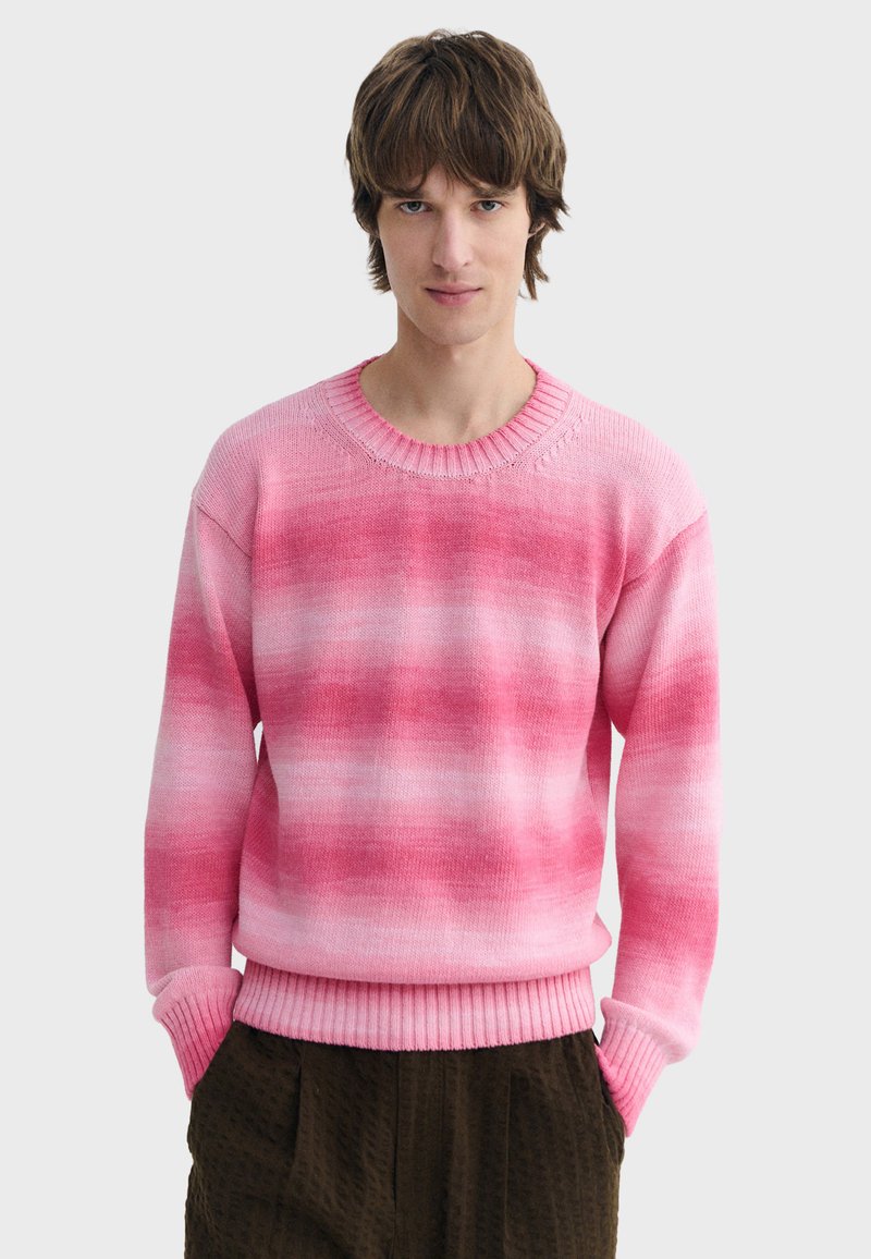 Jeune homme portant un pull en tricot dégradé rose et blanc avec des poignets et un ourlet côtelés, associé à un pantalon texturé marron foncé.