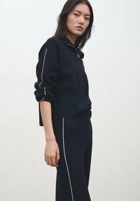 Temno modra pletena majica z visokim ovratnikom in zadrgo, z belo obrobo ob rokavih, združena z ujemajočimi se oprijetimi hlačami.