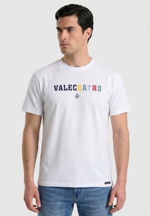 Hombre con cabello oscuro que lleva una camiseta blanca con texto multicolor "VALECUATRO" y vaqueros azules, mirando hacia su izquierda.
