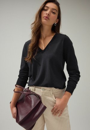 Pulover negru cu mânecă lungă și decolteu în V, fabricat dintr-un material moale, asortat cu pantaloni crem cu dungi subtiri. Într-o mână este ținută o geantă din piele bordeaux închis.