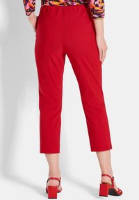 Frau in roten, cropped Hosen, roten Pumps und einem bunten, gemusterten Oberteil, steht mit dem Rücken zur Kamera.