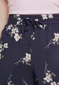 Pantalons à imprimé floral marine avec une taille élastique et un cordon de serrage, en tissu lisse, ornés de fleurs blanches et de feuilles vertes.
