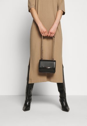 Vrouw in een beige midi-jurk en zwarte kniehoge laarzen, die een kleine zwarte lederen handtas met gouden kettingriemen vasthoudt.