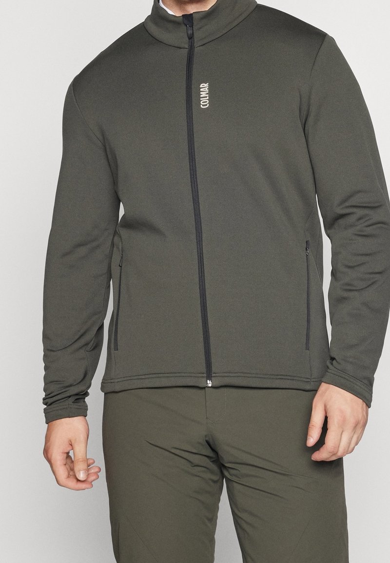 Homme portant une veste zippée vert olive et un pantalon assorti, avec le logo « Colmar » sur la poitrine et des poches latérales zippées visibles.