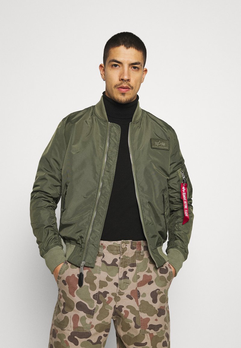 Giacca bomber verde oliva con zip frontale, polsini a costine e toppa sul petto. Presenta un'etichetta rossa sulla manica. Indossata su un dolcevita nero.