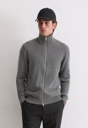 Grauer Reißverschluss-Pullover mit hohem Kragen, aus weichem Material. Hat lange Ärmel und gerippte Bündchen; das Modell trägt dunkle Hosen und eine schwarze Mütze.