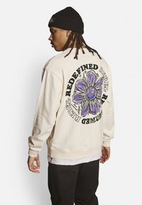 Sudadera crema con un gran gráfico de una flor morada en la parte posterior, que presenta el texto "REDEFINED REBEL". Con puños y dobladillo acanalados, combinada con pantalones negros.