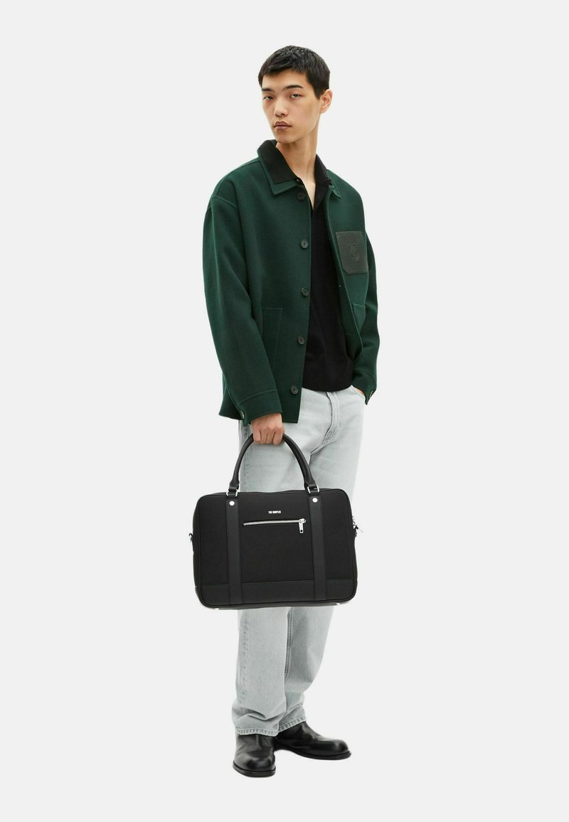 Ung mand iført en mørkegrøn jakke og lyse jeans, der holder en sort aktentaske med en frontlomme med lynlås, stående på hvid baggrund.